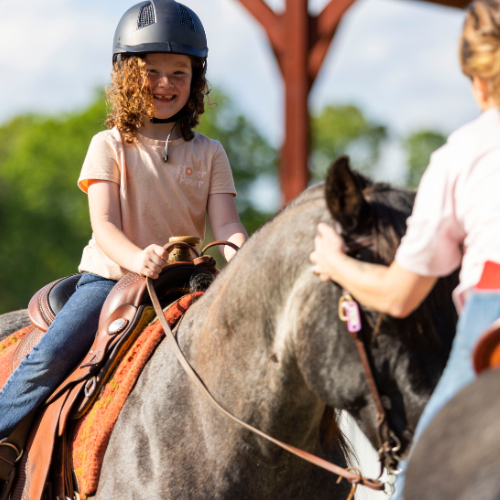 A smiling girl with curly hair wearing a helmet rides a horse guided by a woman. The scene is outdoors with a sunny and joyful atmosphere.