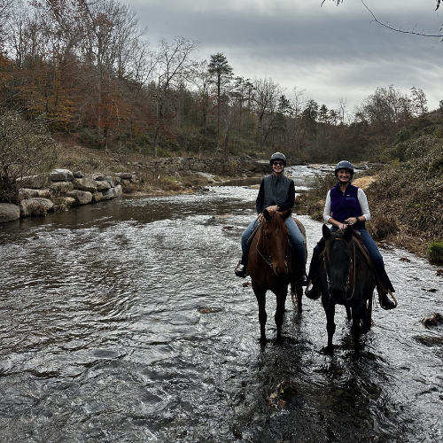 Two people riding horses through a shallow stream, surrounded by autumn foliage and rocks under a cloudy sky, conveying a sense of adventure.