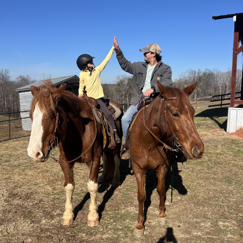 Two people on horses high-five under a clear blue sky. The scene is joyful, with a rustic background and a barn, conveying a sense of adventure.
